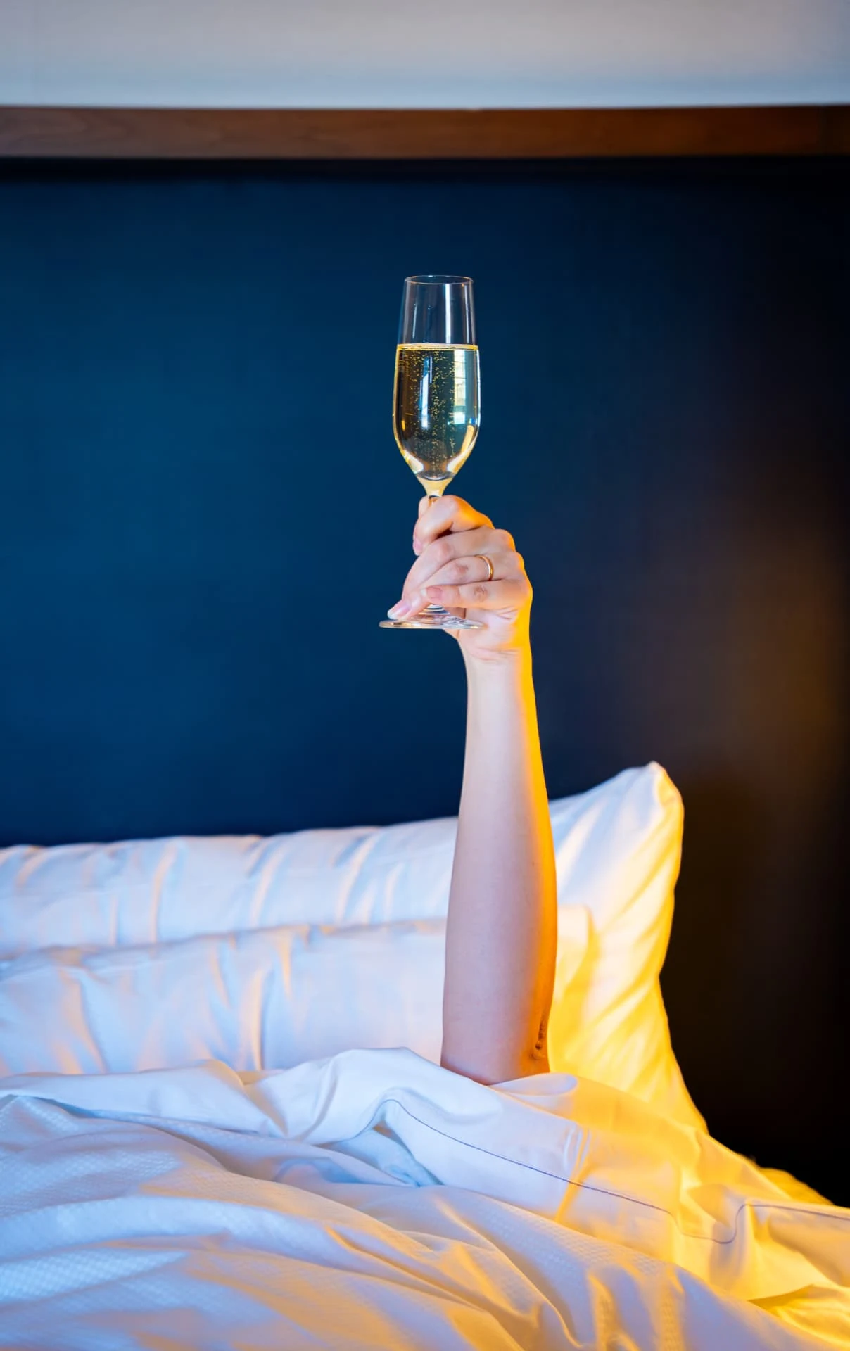 Hand raising a glass of champagne from crisp hotel bed sheets